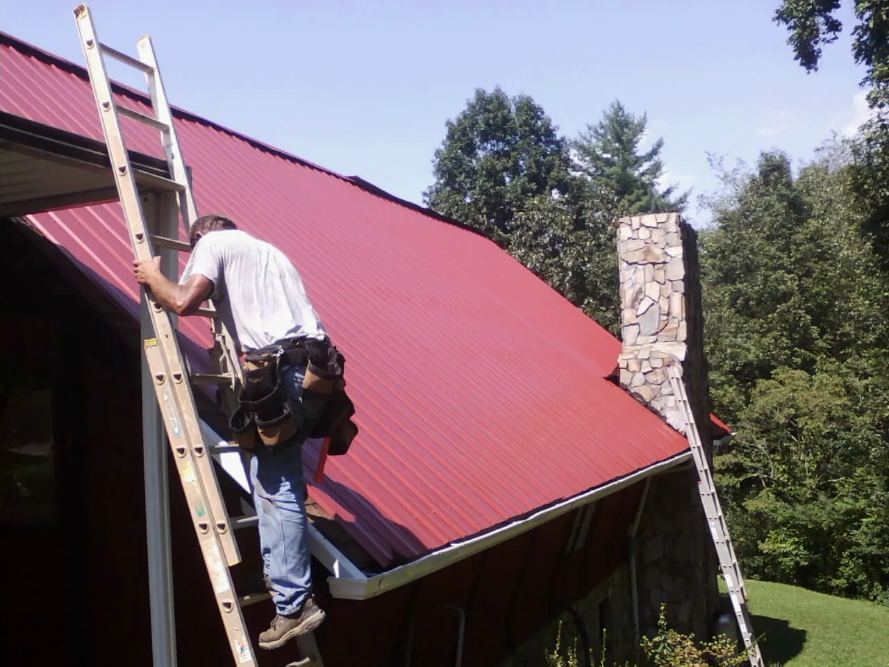 Roofer on a ladder beside a red metal roof and stone chimney — Soffit & Fascia Repair by Roofing Brothers in Brooks