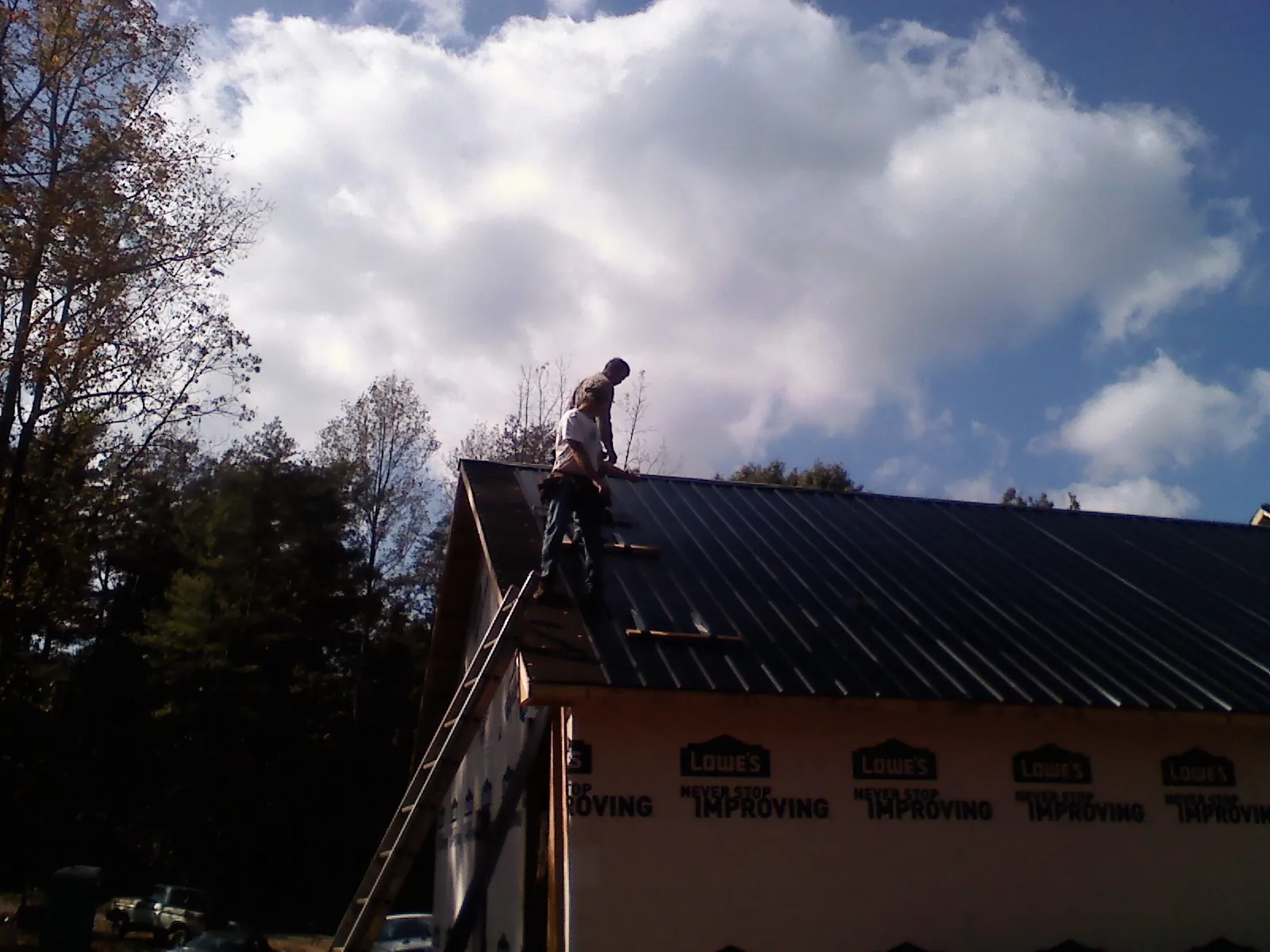 Roofer fastening dark metal panels high against a cloudy sky, from a Roof Inspection job in Brooks